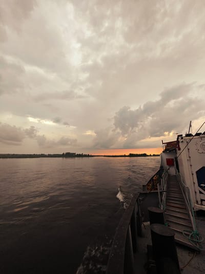 A wide-angle, low-angle, vertical shot captures a river scene from the deck of a boat near Kazachinskoye, Russia. The sky is filled with overcast, grey clouds, with a sliver of orange light visible on the horizon, suggesting either sunrise or sunset. The river stretches across the frame, its surface rippling with dark, choppy water. On the far bank, a line of trees and vegetation forms a dark silhouette against the muted sky. The starboard side of a white vessel occupies the right portion of the image. A metal gangway with steps extends upwards along the side of the ship, and various industrial elements, including cleats and ropes, are visible on the deck. The boat appears to be in motion, with a small wake trailing behind it. The overall atmosphere is somber and overcast.