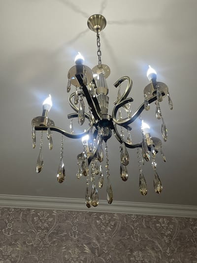 A chandelier hangs from a white ceiling. The chandelier is bronze and black with many clear teardrop-shaped crystals. The bulbs are lit, casting a bright white light. To the right of the image, there is a wall with a floral wallpaper pattern in gray tones. The location is not discernible from the image.