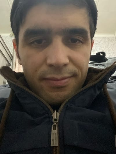 A male individual is depicted in a close-up, head-and-shoulders shot, looking directly at the camera. He has dark hair, pronounced eyebrows, and visible stubble on his jawline and chin. His mouth corners are slightly upturned. He wears a dark blue padded jacket with a brown, possibly faux-fur or fleece, lining visible around the collar. The jacket is partially zipped, exposing a metallic zipper pull with a rectangular, multi-paneled design. The background suggests an indoor setting. To the left, a light-colored wooden wardrobe or cabinet is partially visible. The wall behind the subject is light-colored and features a subtle, possibly patterned, wallpaper. A small, light-colored fixture is visible on the upper left portion of the background wall. The location is in the city of Akhtachi, Uzbekistan.