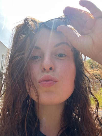 A close-up portrait, captured in Bad Sobernheim, Germany, features a single female subject with fair skin, blue eyes, and long, wavy brown hair. Her lips are pursed in a "kiss" gesture, and she looks directly at the camera. Her right hand is elevated, holding dark-framed sunglasses positioned atop her head. The scene is brightly lit by natural sunlight, causing visible lens flares. The background environment consists of a light-colored building with a visible window on the left, and green foliage against a clear blue sky on the right.