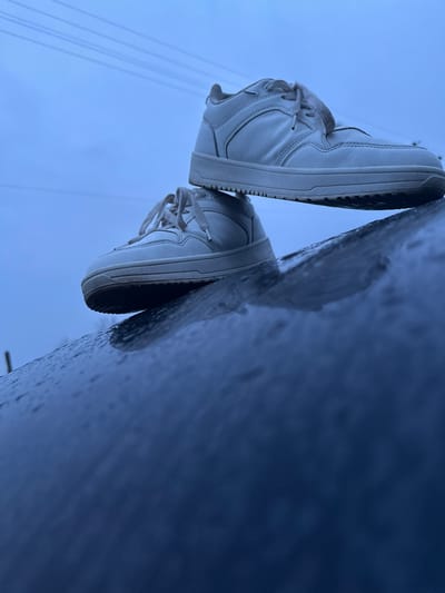 The image displays two white, low-top sneakers stacked on a dark, curved, and wet surface, likely a railing or pipe, in Novgrad, Bulgaria. The surface shows a reflective sheen and numerous small water droplets. The lower sneaker rests directly on this surface, with its sole facing downwards. The second sneaker is positioned on top of the first, oriented similarly. Both sneakers feature visible shoelaces and exhibit minor dirt accumulation consistent with outdoor use. The background consists of a uniformly overcast, light blue sky, against which several faint horizontal utility lines are discernible in the upper portion of the frame. A slender, dark vertical object, possibly a distant pole or tree branch, is partially visible on the far left edge of the horizon. The lighting is diffuse and cool-toned. No individuals are present, and no actions beyond the static arrangement of the footwear are depicted.