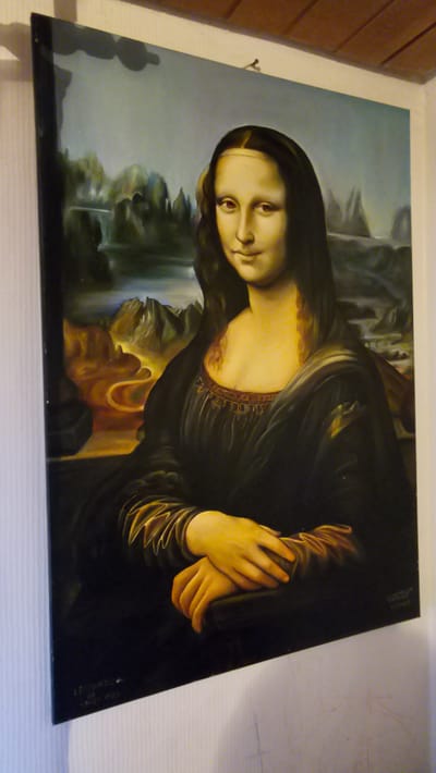 This is a medium shot, eye-level photograph of a painting of the Mona Lisa hanging on a white wall in what appears to be an indoor setting. The painting is centered in the frame and is slightly tilted. The Mona Lisa is depicted with her characteristic enigmatic smile and is wearing a dark dress with intricate detailing around the neckline. Her hands are clasped in her lap, and she is set against a landscape background with mountains and a body of water. There are faint markings on the wall below the painting, possibly from a sketch or writing. The top of the image shows a wooden ceiling with visible planks. The overall lighting suggests an interior space. This image was taken in Oelsberg, Germany.