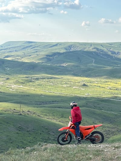 A landscape image depicting a motorcyclist parked atop a grassy incline within the Орджоникидзевский поселковый совет, Ukraine. The rider, clad in a red jacket, blue trousers, and helmet, is seated on an orange dirt bike. The setting is a mountainous terrain with a bright sky visible on the left, contrasting with the grassy slopes of the valley. The scene has a serene, natural atmosphere.