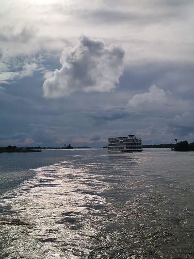 A wide shot captures a cruise ship on a body of water under a cloudy sky. The sun is peeking through the clouds, creating bright reflections on the water's surface in the foreground. The cruise ship, which appears to be a multi-decked vessel with a white superstructure and dark windows, is positioned in the mid-ground, moving away from the viewer. In the distance, the shoreline is visible, with trees and structures. The sky is filled with dramatic, dark clouds, with some lighter, brighter clouds scattered throughout. The overall impression is one of a dynamic and somewhat moody atmosphere on the water. The location is identified as Kargulino, Russia.