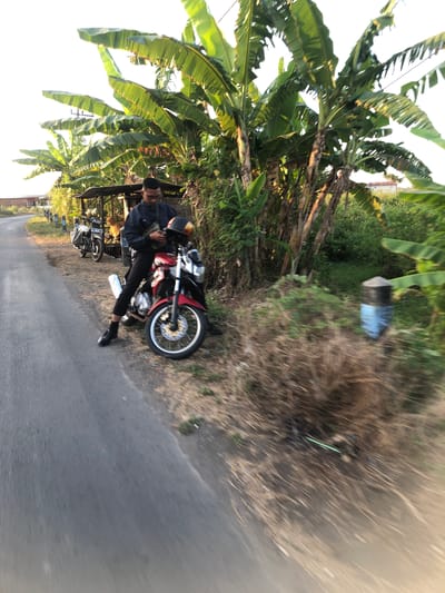 In Glagah Arum, Indonesia, a young man with dark hair and glasses is seated on a red motorcycle. He is wearing a dark blue jacket, black pants, and black shoes. The motorcycle is parked on the side of a paved road, with a grassy verge and banana trees in the background. To the left of the man's motorcycle, another motorcycle is parked near a small wooden structure that appears to be a roadside stall. The sky is bright, suggesting it is daytime. The overall scene depicts a brief stop on a rural road.