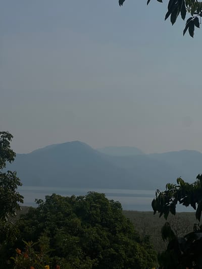 The image shows a hazy landscape in Santa Cruz de Yojoa, Honduras. The view includes mountains obscured by mist, with a body of water visible between them. The foreground is framed by foliage, including trees and bushes, suggesting a viewpoint from a higher elevation.