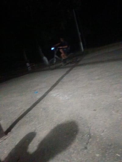 The image depicts a dimly lit outdoor scene, possibly at night, with a person riding a bicycle. The person is visible in the upper-middle portion of the frame, appearing to be on a bicycle with a bright blue light emanating from its front. The ground is a rough, gray surface, possibly asphalt or concrete, with a distinct shadow cast across it from the left side of the frame. In the background, there are dark, indistinct shapes that could be trees or other vegetation, as well as a vertical pole-like structure on the right. The overall quality of the image is poor, with significant blurriness and low light conditions, making precise identification of objects and details challenging. There is no indication of the specific location within Leontyevskoye, Russia, beyond it being an outdoor area.
