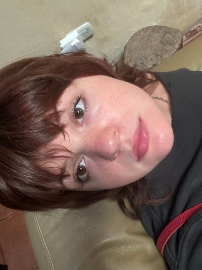 A female subject with shoulder-length reddish-brown hair and bangs is depicted in a close-up, angled portrait, looking directly into the camera. Her fair skin features visible freckles and a slight pink blush on her cheeks. She has brown eyes with mascara applied to both upper and lower lashes, and a silver septum piercing. Her lips are tinted light pink and slightly parted. She wears a dark long-sleeved top and a thin silver chain necklace. A red strap, indicative of a bag or garment, is visible over her right shoulder. The subject appears to be reclining on a light tan, possibly faux leather, upholstered surface, which occupies the lower portion of the frame. In the blurred background, behind and above her head, a white USB wall adapter and an irregular, textured brown object, possibly a piece of stone or wood, are present on a light-colored surface. The scene is located in Antigua Guatemala, Guatemala.