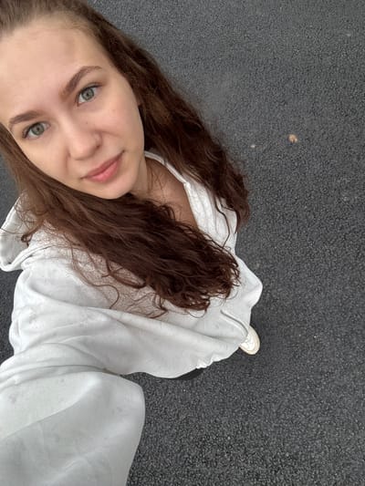 A selfie shot from an overhead perspective depicts a young woman standing on an asphalt surface. She has fair skin, light-colored eyes, and long brown curly hair. The woman is wearing a white hooded jacket. Her face is turned towards the camera. A single white sneaker is partially visible on the right side of the frame. The setting appears to be an outdoor location. The image was taken in the city of Михайловка, Russia.