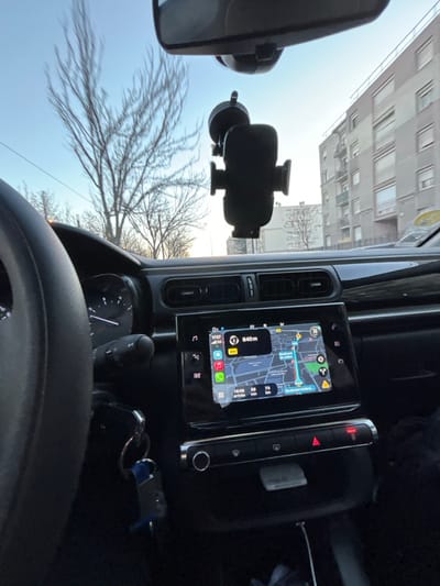 Captured from inside a car, the dashboard dominates the view. A navigation screen displays a map, positioned adjacent to a vertical control panel with buttons. Below, the gear stick and car keys are visible, with the steering wheel in the foreground. A phone mount adheres to the windshield, framing an exterior view of bare trees and buildings against a bright sky.
