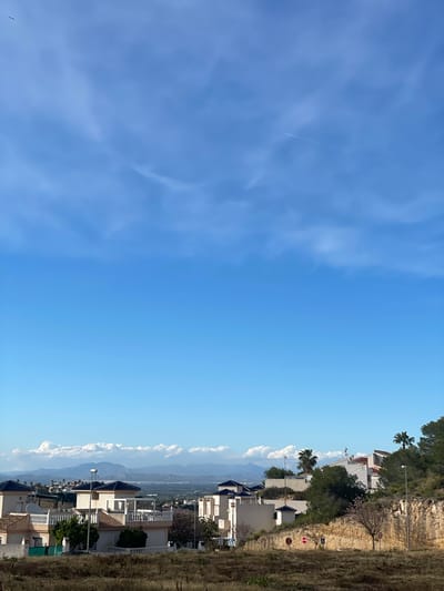 The image shows a view in Rojales, Spain. A residential area with multi-story buildings and some palm trees is visible on the right side of the frame. Beyond the buildings, there is a mountain range partially obscured by low-lying clouds. In the foreground is a grassy area and a stone wall. The left side of the image is dominated by a clear blue sky with some wispy clouds and a contrail. A bird is also visible in the lower left corner.