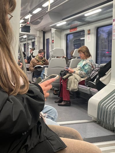 The scene depicts the interior of a train or subway car. Passengers occupy the seats, including a woman in a patterned top using her phone. The focus is on a person in the foreground, partially obscured by long hair and a jacket, who also holds a phone. Seats are visible in the background, along with the train's interior features like windows and overhead lighting. The composition suggests a casual, candid snapshot of everyday life on public transportation.
