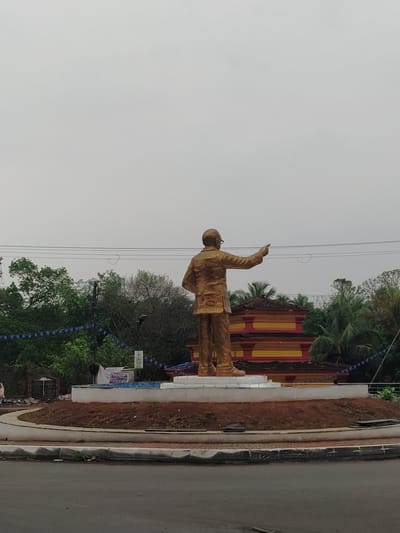 A slightly tilted, medium shot depicts a golden-colored statue of a man wearing glasses and a suit, with his right arm raised pointing upwards. The statue is situated on a platform with a red-brown dirt border alongside a road. The background includes a structure with red and yellow detailing, trees, and power lines against a cloudy sky. This is a statue of B. R. Ambedkar in Chopdem, India.