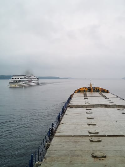 This is an aerial view of two vessels on a body of water, possibly a river or lake, under an overcast sky. In the distance to the left, a white multi-decked passenger ship is seen moving away from the camera, emitting a plume of dark smoke from its stack. The ship has multiple windows and balconies. In the foreground and occupying the right side of the frame is the deck of a large, flat-topped barge or cargo ship. Its deck is concrete, with numerous rectangular hatches or manholes. The barge has a blue railing along its left side. The water is calm with some ripples. The background shows a distant treeline on the horizon. The location is specified as the city of Khoroshevo, Russia, implying this is a scene on a waterway within or near the city. There are no people visible in the image.