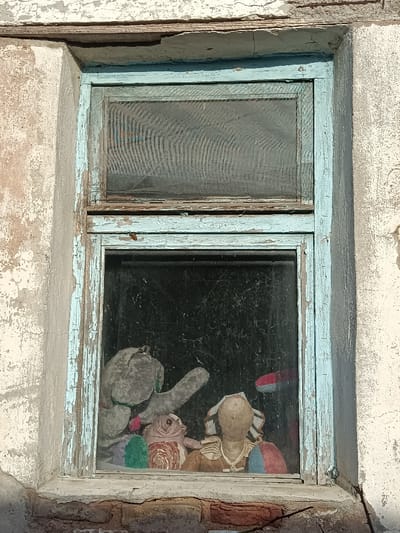 A weathered window frame, painted a faded blue, shows significant chipping and peeling. Behind the dusty glass, a collection of well-worn stuffed animals is visible. They appear huddled together, their colors muted and their forms softened with age. The window is set into a wall with peeling paint. The scene evokes a sense of abandonment and nostalgia.