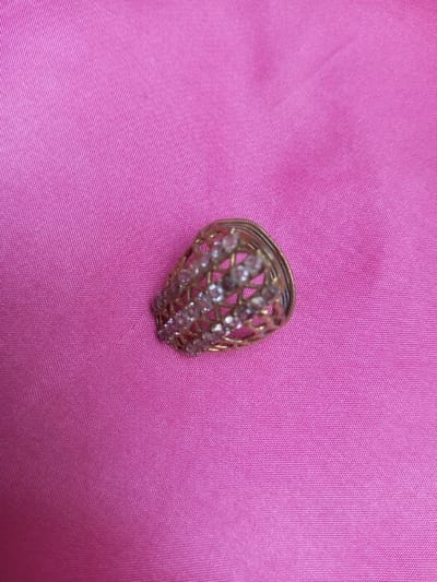 A close-up image, taken in Qarabucaq qəsəbə, Azerbaijan, displays a gold-colored ring with an ornate, lattice-style band encrusted with multiple rows of small, clear, faceted stones. The ring is positioned on its side, resting on a vibrant pink fabric surface. The fabric exhibits a fine weave texture and subtle undulations. No individuals are present, and no specific actions or interactions are discernible.
