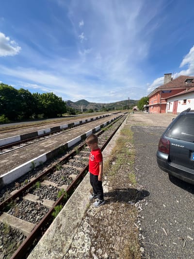 A young boy in a red t-shirt and black pants stands on a train platform in Stareishino, Bulgaria. The platform has train tracks next to it, and a dark-colored car is parked on the asphalt next to the platform. In the background, there is a train station building with a sign that reads "PODKOVA PODKOVA". Mountains and trees are visible in the distance under a blue sky with white clouds.