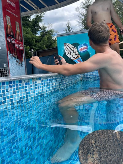 This is a horizontal, eye-level medium shot of two young men in a swimming pool in Dobrinishte, Bulgaria. The man in the foreground, partially submerged in the bright blue, tiled pool, is leaning over the edge, looking at his phone, which he holds in his right hand. He wears patterned swim trunks. The second man, to his left and further in the background, is also in the pool, wearing orange swim trunks with a pineapple pattern.

Behind the men, a light blue cooler or vending machine displays an image of a Nestle ice cream cone and the words "ICE CREAM" and the Nestle logo. To the left of this, a red sign with "Coca-Cola" logos and the tagline "TASTE THE FEELING" is visible. The background features lush green trees and a cloudy sky, suggesting it is daytime. The water in the pool is clear and reflects the surroundings. The overall impression is one of casual relaxation on a warm day.