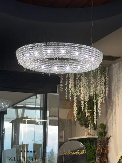 The image shows an ornate crystal chandelier hanging from a dark ceiling. Below the chandelier, to the right, is a decorative display featuring cascading white artificial flowers, a golden candelabra, and lush greenery. In the foreground, a circular black sign with golden cursive text reads "Dobrodošli". To the left, a glass door with "HOTEL HILLS" and three stars is visible, suggesting the location is Hotel Hills in Ilidža, Bosnia and Herzegovina. The environment appears to be an indoor space, possibly a hotel lobby or reception area.