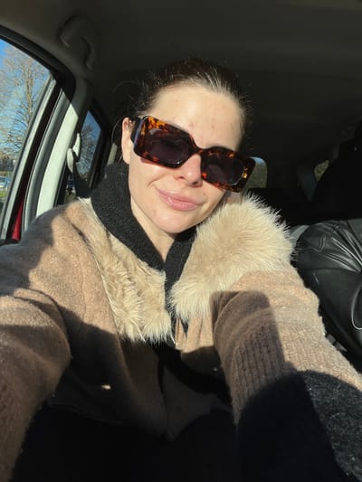 A female subject is depicted in a close-up, front-facing selfie taken within the interior of a vehicle. She occupies the driver's seat, partially obscuring the car's interior. The subject wears large, rectangular, tortoiseshell-patterned sunglasses. Her hair is pulled back. She presents a slight smile with visible gloss on her lips. Her skin exhibits multiple small, red blemishes across the forehead and cheeks. She is dressed in a black hooded garment layered beneath a light brown, possibly knitted or woolen, jacket featuring a prominent beige faux fur trim around the collar and lapel area. Her left arm, clothed in the same light brown material, is extended to hold the camera. Strong sunlight enters the vehicle from the left, creating distinct shadows on the subject and interior elements. Through the driver's side window, a clear blue sky, bare deciduous trees, and an indistinct, possibly suburban, background are visible. A second individual is partially visible in the passenger seat to the right, clad in a black, leather-like jacket. The specific location within the city of Marquette-lez-Lille, France, cannot be determined from the visual evidence present in the image.