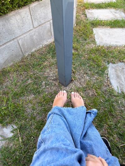 This image, located in Quilmes Oeste, Argentina, displays a close-up, downward-angled view of a person's lower body within an outdoor environment. The subject's bare feet are prominent in the foreground, resting on sparse grass. The toenails on both feet are painted white. The individual is wearing loose-fitting, medium-wash blue denim jeans, which extend from the bottom of the frame to just above the feet. A hand, with visible patterned fingernails, is briefly visible at the bottom right corner, resting on the leg.
The ground consists of a mixture of green and dry grass with visible patches of soil. Centered in the upper half of the frame is a dark grey, rectangular vertical post, approximately three to four inches in width, its base embedded in the ground. To the left of the post, a low wall constructed of light grey concrete blocks is visible, topped by dense green foliage, likely a hedge. To the right of the post, a pathway composed of light grey square concrete pavers is partially visible, interspersed with grass.