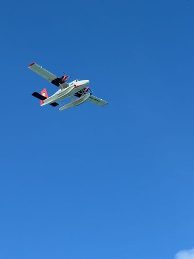 An aerial view captures a white and red seaplane flying against a clear blue sky. The seaplane, identified by the registration "8Q-MAJ," has two propellers and floats beneath its fuselage. The plane is angled upwards, with its left wing higher than its right. The tail of the aircraft features a red and black insignia. The scene is devoid of any immediate environmental context like clouds or landmasses, suggesting an unobstructed view of the sky. The image appears to be taken from a lower vantage point, looking up at the seaplane. The registration "8Q-MAJ" indicates the seaplane is registered in the Maldives, and Malé, as the capital and a major hub for seaplane operations in the Maldives, is a likely location for such a scene.