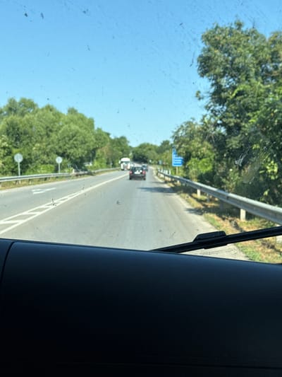 The image depicts a view through a car windshield on a clear, sunny day. The road ahead is paved with white lane markings. A dark-colored car is in front, and a white truck is further down the road. Trees line the sides of the road, and a metal guardrail runs alongside it. A blue sign is visible in the distance. The foreground of the image shows the dark dashboard of the vehicle.
While the image itself does not explicitly confirm the location as Osenets, Bulgaria, the presence of road signs and guardrails suggests a roadway in a populated area. Further information would be needed to definitively pinpoint the location within Osenets.