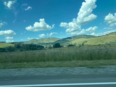 The image captures a scenic view of rolling green hills under a vibrant blue sky dotted with fluffy white clouds. The foreground is dominated by a blurred expanse of tall green grass, suggesting the photo was taken from a moving vehicle. Beyond the grass, a line of trees marks the transition to the hills, which are covered in lush vegetation and appear to be uninhabited. There are no visible people or structures in this landscape, creating a sense of natural tranquility. The image appears to be taken during daylight hours. While the location is stated to be Verkh-Aya, Russia, there are no specific landmarks or details within the image to confirm this or any other specific geographical location.
