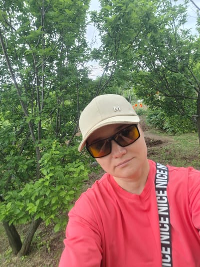 In Svobodny, Russia, a woman wearing a coral-colored t-shirt, a beige baseball cap with an "M" logo, and dark sunglasses poses for a selfie in a natural outdoor setting. She is positioned on a dirt path surrounded by lush green trees and foliage. The strap of a bag or camera is visible across her chest, with the repeated word "NICE" printed vertically. The background shows more trees, a slightly elevated path, and hints of orange flowers in the distance. The lighting suggests an overcast day.