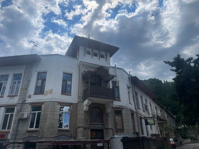 A low-angle, wide shot captures an old, white and stone-clad building in Livadiyskyi settlement council, Ukraine. The building has a detailed architectural style with multiple windows, some reflecting the cloudy sky. A small balcony with ornate metalwork is featured prominently on the facade. A weather vane is visible on the roof. The sky is filled with dramatic, dark clouds interspersed with patches of blue sky. Lush green trees rise behind the building, suggesting a hillside location. The street in front of the building appears to be paved, and a small red car is parked to the right. The overall impression is one of historical architecture set against a dynamic sky.