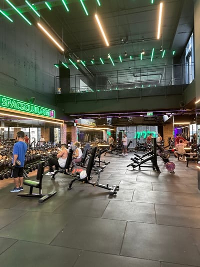 This image depicts the interior of a modern gym, identified by a prominent green neon sign as "SPACECLUB.GYM," located in Beer Yaaqov, Israel. The facility features a high, dark gray ceiling equipped with numerous linear green LED lights and several warm yellow/white tube lights. A mezzanine level with a black metal grid railing is visible in the upper mid-ground, also illuminated by green neon strips.

The main gym floor is covered with large, rectangular dark gray rubber mats. The walls are a mix of dark gray paint, exposed brick patterns, and large mirrored sections that reflect the gym's interior and lighting.

Approximately ten to fifteen individuals, primarily adults, are present. In the foreground left, a male in a blue t-shirt and dark shorts stands, holding a dumbbell in his right hand, facing away from the camera, next to a black adjustable weight bench. He wears headphones. To his right, in the mid-ground, a male in a white tank top and black pants is seated on an adjustable weight bench, looking down at a mobile device. Adjacent to him, a female with light, curly hair, wearing a white top and dark pants, is seated on another adjustable bench, facing towards the right side of the frame.

The gym floor is populated with various black gym equipment, including multiple adjustable weight benches, dumbbell racks lining the left wall, and an assortment of weight machines and cable equipment in the mid-ground and background. Purple barbell plates are visible on some machines. Several other individuals are dispersed throughout the space, engaged in activities such as using weight machines, lifting free weights, or standing and observing.