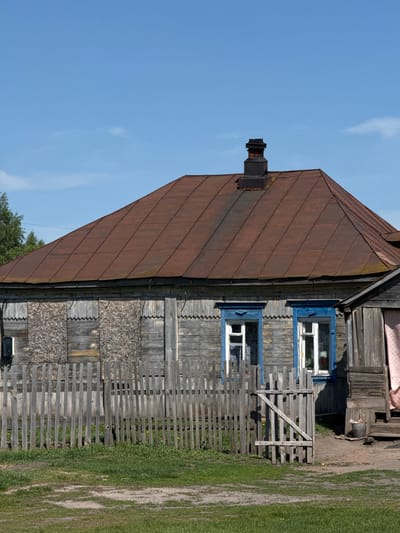 The image shows the side of a rustic wooden house with a rusty brown metal roof. The house has two visible windows with bright blue frames. A wooden fence runs along the side of the house where it meets the grassy ground. The sky is blue with faint white clouds. The house is located in the city of Слобода, Russia.