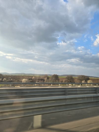 The scene depicts a rural landscape observed from a moving vehicle on a road or highway near Malko Tranovo, Bulgaria. In the immediate foreground, a three-tiered grey metal guardrail extends horizontally across the frame, exhibiting motion blur consistent with vehicular movement. Beyond the guardrail, the midground features cultivated fields of brown soil interspersed with patches of green vegetation and numerous deciduous trees; some trees are bare, while others are covered in white blossoms. The background reveals a line of green, rolling hills under a sky dominated by large, light grey and darker cumulus clouds, with visible patches of blue sky, particularly towards the upper right. Sunlight penetrates the cloud cover, illuminating the left side of the scene. No individuals, specific actions, or interactions are present.