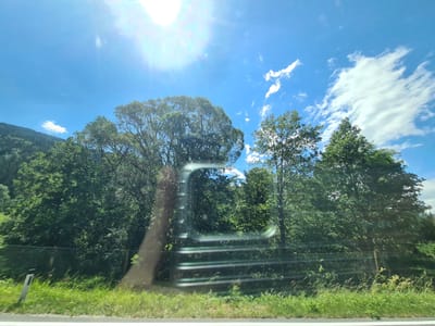 A low-angle, outdoor, wide shot captures a roadside scene on a bright, sunny day, likely taken from a moving vehicle. The foreground features a grassy verge next to a paved surface, with a white marker post on the left. A chain-link fence runs horizontally across the middle ground, separating the road from a dense line of lush green trees. The trees form a thick canopy, obscuring the terrain behind them. In the upper portion of the frame, a bright sun flares in the upper left corner, casting a lens flare across the image. The sky is a vibrant blue, dotted with scattered white clouds. The overall impression is one of a rural or semi-rural landscape. The location is Wildbad Einöd, Austria.