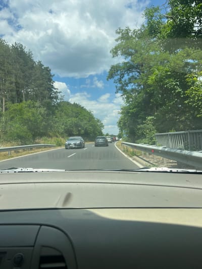 The image shows a view from inside a car driving on a highway. The highway is lined with trees and dense vegetation on both sides. The sky is partly cloudy with patches of blue visible. There are several cars on the highway in front of the vehicle. The dashboard of the car is visible in the foreground. The scene appears to be a rural or suburban area, given the abundant greenery. The location is identified as the city of Balabanovo, Bulgaria, though the image itself does not provide explicit visual cues to confirm this specific location, such as road signs or distinctive landmarks.