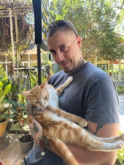 A man with dark hair and tattoos is holding an orange and white cat. He's wearing a gray shirt and sunglasses are perched on his head. The cat is sprawled out in his arms. The scene appears to be outdoors, with trees and plants in the background. A sign with writing is visible to the left. The man is looking at the camera with a slight smile.