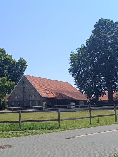 A long, traditional brick building with a red-tiled roof stands under a clear blue sky. The building has several windows and a large gable. In front of the building is a wooden fence bordering a grassy field. A large tree is visible to the right of the building. The foreground shows a paved walkway.