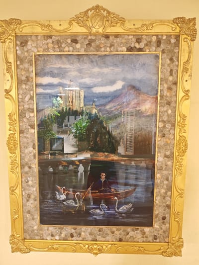 This is a photograph of a framed painting. The painting depicts a scene on a body of water, with a castle-like structure in the background. In the foreground, a person is rowing a boat on the water, accompanied by several swans. The frame of the painting is ornate and gold-colored, and it is decorated with small stones embedded in it. The location in the city of Schwangau, Germany is not discernable from the image itself.