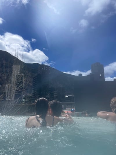 A vertically-oriented image captures a scene in Les Escaldes, Andorra, possibly from the Caldea thermal spa. The foreground shows figures waist-deep in water, with their backs to the camera. In the background, a large building is visible, set against a mountain backdrop. The sky is blue with scattered clouds. Bare tree branches are also present.