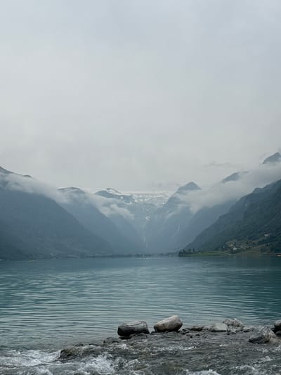 The image depicts a serene natural landscape. In the foreground, a rocky shore meets the calm, turquoise waters of a lake. The water's surface is rippled, suggesting a gentle breeze. Several large, grey stones are scattered along the shoreline.
Beyond the lake, majestic mountains rise steeply, their slopes covered in lush green vegetation. A notable feature of the mountains is a visible glacier situated at a higher elevation, partially obscured by swirling clouds that also hug the mountain peaks. The sky is overcast and a pale grey, contributing to the moody atmosphere of the scene.
On the distant shores of the lake, nestled at the base of the mountains, small clusters of houses are visible, hinting at a human presence in this otherwise wild environment. The overall impression is one of a remote, tranquil, and awe-inspiring natural setting in the vicinity of Olden, Norway, a region renowned for its dramatic fjords and glacial landscapes.