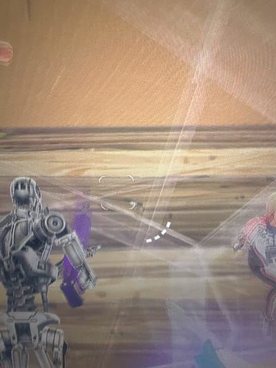 This is a screenshot from a video game. It depicts two robotic characters in a sandy, desert-like environment. The character on the left is a humanoid robot with a metallic gray finish, a detailed chest piece, and what appears to be a weapon or tool in its left hand. The robot on the right is partially visible and appears to be wearing red and white armor. There are glowing, translucent lines and target reticles superimposed on the image, indicating gameplay elements. The background is a textured, sandy expanse with horizontal bands of color that suggest distance or terrain features. There is no indication of a city or any specific location like Bezden, Bulgaria.
