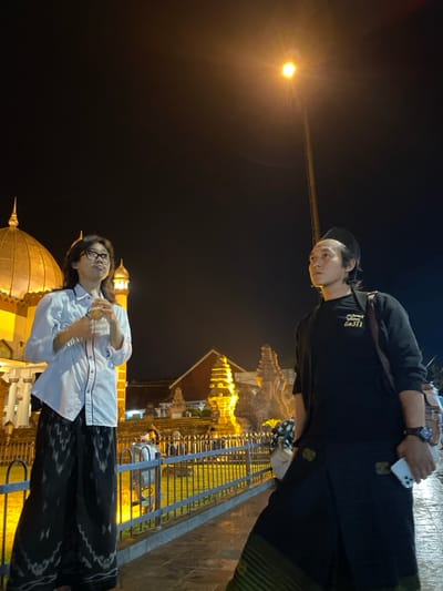 At night in Kauman, Indonesia, two adult males stand on a paved surface, illuminated by a prominent yellow-orange streetlamp. To the left, a male with long dark hair and glasses wears a white long-sleeved shirt with dark trim and dark patterned sarong-style trousers; he holds a translucent object at chest level and looks upward-left. To the right, another male wears a black peci, a black long-sleeved shirt with a light emblem, dark shoulder bag, and dark sarong-style trousers with a green patterned lower band; he holds a white smartphone in his left hand and looks upward-right. In the background, behind a dark metal fence, are brightly lit architectural structures: to the left, golden domes and a minaret-like structure indicative of a mosque; to the right, intricately carved stone structures with tiered roofs, consistent with traditional Javanese architecture.