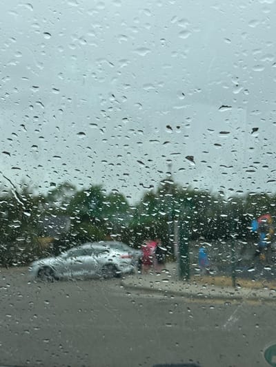 The image depicts a view through a rain-streaked car window, looking out onto a street scene in Courtown, Ireland. The sky is overcast and the weather appears to be rainy. In the mid-ground, a silver car is visible, parked or driving slowly on the asphalt road. Behind the car, a line of trees forms the backdrop. Further in the background, the indistinct shapes of buildings and possibly a traffic light or signal can be seen, with flashes of color, one appearing red and another blue. The foreground is dominated by numerous raindrops clinging to the car's window, distorting the view of the outside. The overall impression is one of a damp and gray day in a suburban or town setting in Courtown.