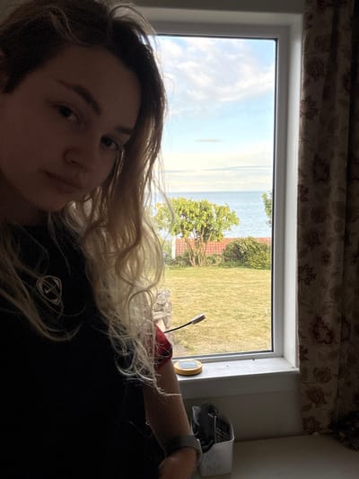 A young woman with long, blonde hair, wearing a black shirt with a red and white logo, sits by a window looking out at the view. The window is open, and she is looking out at the sea in the distance. The view from the window shows a small garden with green grass, trees, and a brick wall. There are some objects sitting on the window sill. One is a small, solar-powered light. The woman is wearing a silver bracelet on her wrist. This appears to be a residential building in Crail, a coastal town in Fife, Scotland. The building is located near the harbor, overlooking the Firth of Forth. The woman is looking out at the ocean, possibly watching the boats in the harbor or enjoying the sea breeze. The image appears to have been taken from inside a home or an apartment. The woman is seated and appears to be calm and relaxed. There is a small, white container with handwritten writing visible on the windowsill. The view is of the sea and coastline, suggesting that the building is located on a cliff overlooking the water.