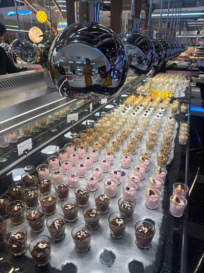An eye-level, full shot showcases a dessert display. Rows of small glass cups containing various treats, like mousse and puddings in colors like brown, pink, and tan, are arranged on a surface marked with circular stains. Silver metal globes reflect the surrounding environment, while a sign that says "Kreme Cafe" and "Vaciar Fruits" is near the desserts.