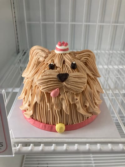 An image taken in Krabi, Thailand, depicts a dog-shaped cake on a white square cake board, resting on a white wire shelf. The cake is primarily light brown, decorated with piped buttercream to simulate fluffy fur on a dog's head. It features two dark brown spherical eyes and a dark brown heart-shaped nose. A small pink oval shape, resembling a tongue, protrudes from the lower left side of the face area. On top of the head, a small, multi-tiered white and pink miniature cake decoration is placed. Around the base of the cake is a pink band, resembling a collar, with a small yellow spherical decoration at the front. The cake board and the wire shelf suggest the item is displayed within a commercial refrigerator or cooler, with additional wire shelving visible in the background. No people are present, and no actions or interactions are occurring. A partial white plastic sign holder is visible on the far left edge of the frame.
