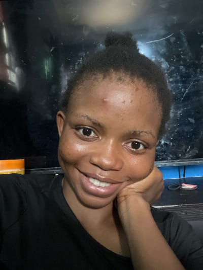 A close-up, rotated selfie of a Black woman with short, dark hair pulled into a small bun. Her right hand is positioned under her chin. She is smiling, showing her teeth. She is wearing a black t-shirt. The background is dark and indistinct, but appears to be a reflective surface. There is no indication of the location being Onibudo, Nigeria in the image.