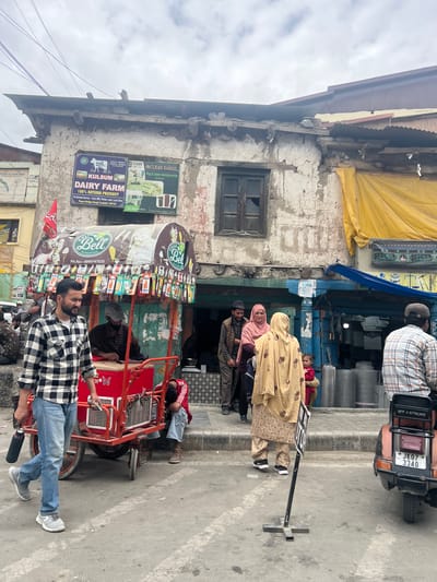 The image depicts a street scene in Kargil, India, on a cloudy day. In the foreground, a man in a black and white checkered shirt and blue jeans walks past a red ice cream cart. The cart is adorned with colorful ice cream packaging and a "Green Bell" logo. Behind the cart, a man in a striped shirt and a cap is seated. Further back, a group of people are gathered near a store with a yellow awning. The store displays various items, including what appear to be construction materials. A scooter with a license plate reading "JK07 3340" is parked on the right side of the image. The buildings in the background show signs of age and wear. A sign above the ice cream cart advertises "KULSUM DAIRY FARM 100% NATURAL PRODUCT".