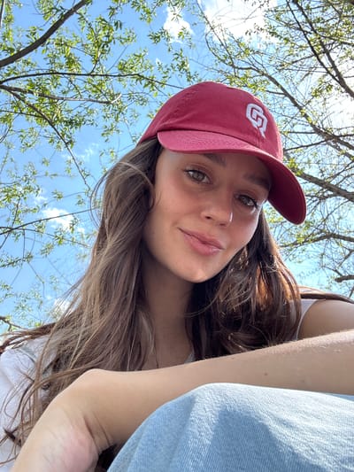 A young woman with fair skin and brown hair is facing the camera. She is wearing a red baseball cap with a white logo on the front. She is wearing a white top and a light blue denim jacket or shirt. The background shows a blue sky with tree branches and young green leaves. The location of the city of Овсянка, Russia cannot be confirmed from the available image.