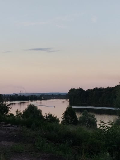 The image captures a serene, dusky scene at a body of water, likely a river, in Mikhailovka, Russia. The sky is a gradient of pale pink and blue, with faint clouds streaking across. In the distance, industrial structures emit plumes of smoke, indicating a nearby industrial zone. A small boat is visible on the water, creating a wake as it moves. The foreground is dominated by dense, dark green foliage, suggesting a natural or park-like setting. The overall atmosphere is peaceful, with the natural elements of the water and trees juxtaposed against the distant industrial landscape.